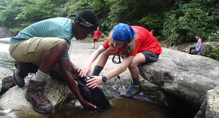 Two students kneel on rocks as they fill a water bladder in shallow water.